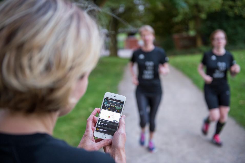Eine Person hält ein Smartphone mit Laufdaten, während zwei Frauen im Hintergrund joggen.